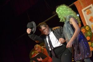aktorzy prezentują scenkę: gruby mężczyzna w okularach zdejmuje kapelusz, rozmawia z partnerką która ma zielone włosy
